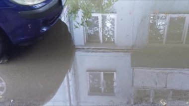 Bir araba yoldaki su birikintisinin içinden geçiyor, ıslak bir yüzey üzerinde sıçramalar ve hareketlilik yaratıyor..