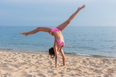 Sahilde bir çocuk spor yapıyor ve denizin arka planında jimnastik yapıyor. Bir çocuk sahilde amuda kalkar.