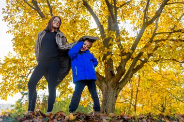 Küçük erkek kardeşiyle birlikte bir kız kardeş sonbahar sarı ağacının yanında duruyor.