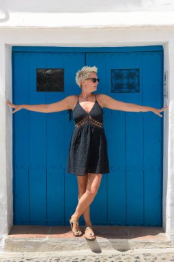 Kısa saçlı, kısa etekli ve güneş gözlüklü güzel bir kadın mavi ahşap kapının yanında poz veriyor..