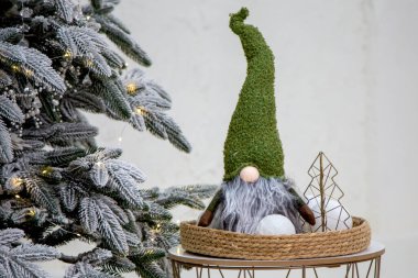 Yeşil desenli şapkalı ve tüylü gri sakallı büyüleyici bir noel cücesi örülmüş bir tepside dekoratif kartopları ve metal tel bir ağaçla birlikte sunuluyor.