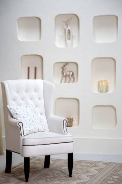 Modern minimalist interior featuring a white tufted armchair with a decorative pillow, set against a wall with built-in niches showcasing elegant holiday decor, including a metallic reindeer and glowing candles clipart