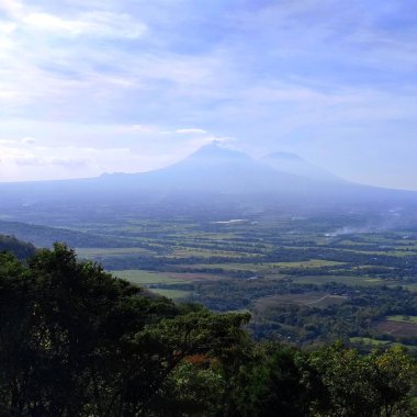Tepenin tepesinden arka planda etkin bir volkan ve bulutlu mavi bir gökyüzü ile birlikte kara manzarası