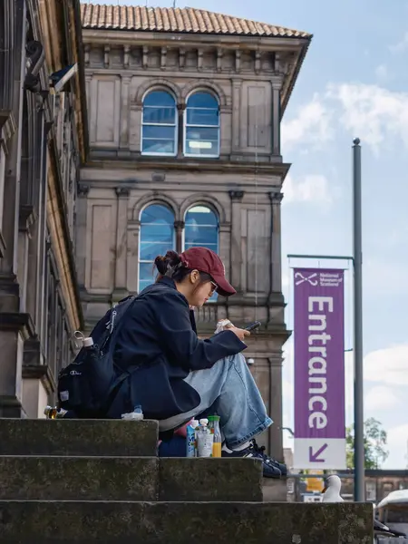 EDINBURGH - 28 Temmuz 2024: Genç bir Asyalı Edinburgh Ulusal Müzesi 'nin merdivenlerinde oturmuş, atıştırmanın tadını çıkarıyor ve akıllı telefonlara bakıyor..