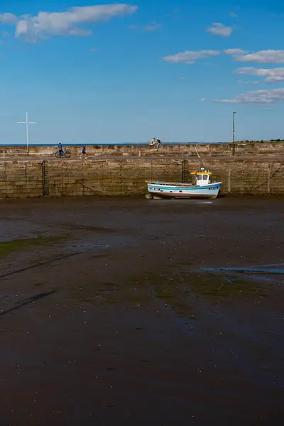 stock image EDINBURGH - JULY 28, 2024: A small fishing boat is grounded on the mudflats in low tide in Fisherrow Harbour, with a clear blue sky and distant figures exploring the shore in the background.