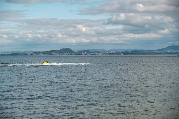 BURNTISLAND, FIFE, SCOTLAND, İngiltere - 11 Haziran 2024: Serin su üzerinde canlı sarı bir jet ski hızlanarak, arka planda parçalı bulutlu bir gökyüzünün altında Edinburgh şehrinin ufuk çizgisini gösteriyor.