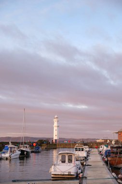 The lighthouse stands tall in Newhaven Harbour in Edinburgh, surrounded by boats as the sun sets, casting warm colors across the water and sky. clipart