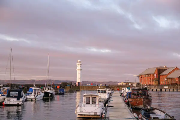 Edinburgh 'daki Newhaven Limanı üzerinde sakin bir günbatımı, deniz fenerini ve demirli tekneleri vurguluyor; sakin sular gökyüzünün sıcak renklerini yansıtıyor.