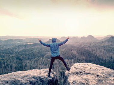 Happy man in blue sweatshirt with raised arms gesture triumph on exposed cliff. Satisfy hiker silhouette on sandstone cliff watching down to hilly landscape. 