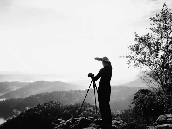Photographer  on cliff. Nature photographer takes photos with mirror camera on peak of rock. Dreamy fogy landscape, spring orange pink misty sunrise in a beautiful valley below.