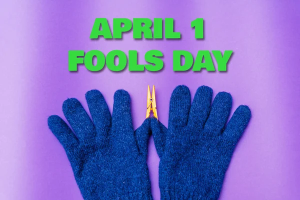 April 1 fools day inscription.A Pair of Knitted blue Gloves on purple Background.The thumb has a Laundry clip.April fool day concept.Copy space.