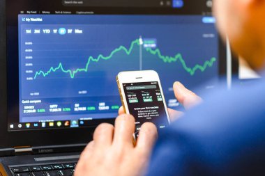 İşadamı akıllı telefonu ve dizüstü bilgisayarı ile borsayı kontrol ediyor. Borsa alım satım verilerini analiz ediyor. Ticaret ve finans konsepti. Telsiai, Litvanya. 03-30-2023