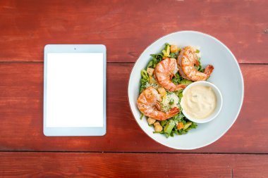 Karides Salatası 'nın yanında boş beyaz masaüstü ekranı olan dijital tablet. Ahşap masa üzerinde karidesli sağlıklı deniz ürünleri Sezar Salatası..