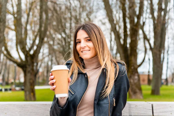 Joyful young woman with coffee paper cup relaxing on bench in autumn park.A woman is holding a brown coffee cup in her hands.Woman in autumn park.Closeup.