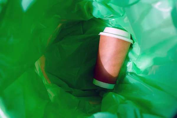A green garbage bag in the trash. There is a paper coffee cup inside.Top view.