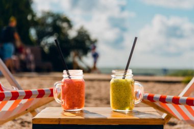 Lüks tropikal sahilde koyu kırmızı ve yeşil renkli iki meyveli kokteyl. Bulanık deniz göğü ve tatilcilerin geçmişi..