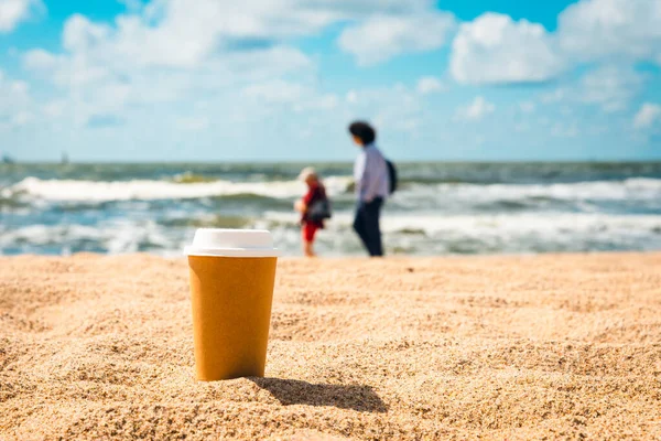 paper cup of coffee, tea stands in the warm sand. On the background of the beautiful sea and the cloudy day.Paper brown cup of coffee on the sand beach over cloudy sky and sea.