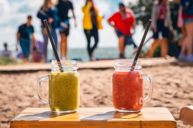 Lüks tropikal plajda koyu kırmızı ve yeşil renkli iki meyveli kokteyl. Bulanık deniz göğü ve tatilcilerin arka planı..
