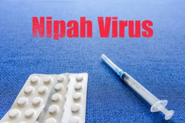 Nipah virüsü kavramı. Üzerinde NIPAH VIRUS yazılı mavi bir arka plan. Üst görünüm, masada duran şırınga hapları..