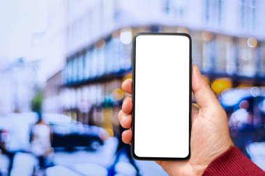 Akıllı telefondan oluşan boş bir beyaz ekran. Logonuz veya tasarımınız için şablon ekranlı modern siyah cep telefonu tutan bir erkek el. Arka planda bulanık bir sokak..