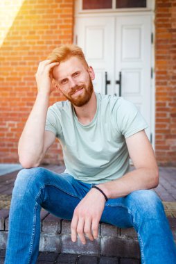 Mutlu Yakışıklı Sakallı Adam, Eski Tuğla Duvar Binası 'nın yanında oturmuş kameraya bakıyor. Açık hava çekimi..