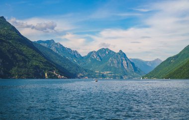 Güneşli bir yaz gününde arka planda yeşil İsviçre Alpleri ile Lugano Gölü boyunca seyreden sürat teknesinin güzel panoramik manzarası. Ticino Kantonu, İsviçre.