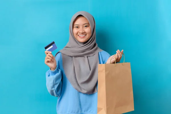 Young Indonesian woman with hijab holding delivery box and a debit/credit card, Isolated on blue background