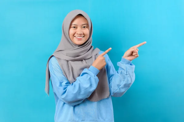 陽気なインドネシアのイスラム教徒の女性は コピースペースに離れてポイントし 素晴らしいプロモーションを議論し 道や方向を与え 青いセーターを着て 楽しい笑顔を持っており 楽観的で 青い背景に隔離された感じ — ストック写真