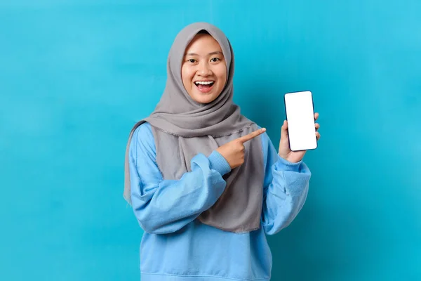 幸せ美しいです若いですインドネシアの女性でヒジャーブは 青い背景に隔離された携帯電話の画面で白い画面の携帯電話と指差し指を示しています — ストック写真