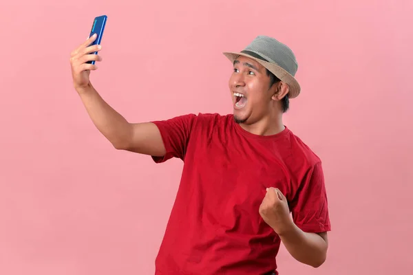 スマートフォンを持っている若いアジアの男は 携帯電話を保持して勝利ジェスチャーを行う 幸せなピンクの背景に隔離された特別な贈り物をオンラインで入手 — ストック写真