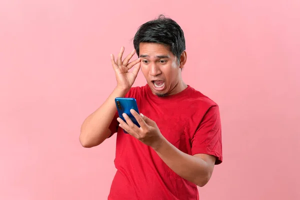 吃惊地看着手里拿着智能手机的亚洲人 眼睛睁得大大的惊讶地看着手机 脸上露出惊讶的表情 孤立的粉色背景 — 图库照片