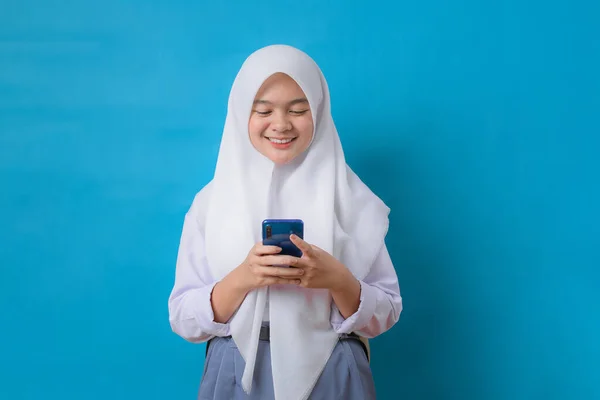 印度尼西亚年轻学生手持带着制服和头巾的智能手机 蓝色背景隔离的微笑 — 图库照片