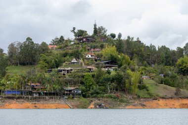 A scenic view of houses situated on a lush hillside by the Guatape Reservoir in Antioquia, Colombia, with trees and water surrounding the area, a popular destination for tourism. clipart