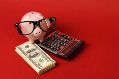 Siyah gözlük çerçeveli domuz kumbarası ve yığınla para üzerinde duruyor. Yüz dolarlık banknotlar ve kırmızı arka planda hesap makinesi.
