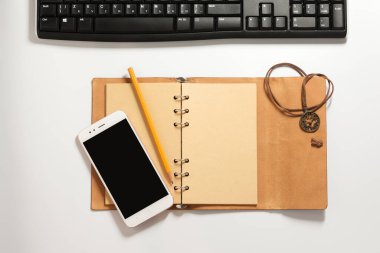 Sarı boş sayfaları olan açık bir defter, güzel bir deri ambalaj, dekoratif dantel tılsım, gri bir kalem, akıllı telefon ve açık gri gradyan arka planda siyah klavye..