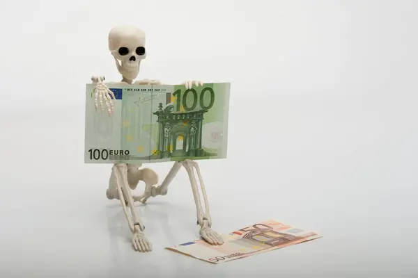 Elinde 100 avroluk banknot tutan ve beyaz arka planda oturan oyuncak bir insan iskeleti.
