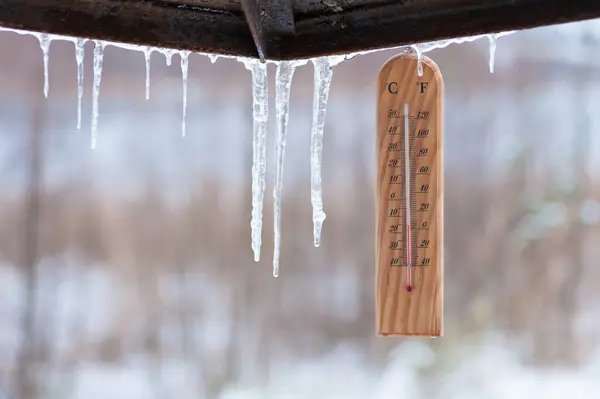 Bir ahşap termometre buz saçaklarının arasında asılı duruyor ve hava sıcaklığını gösteriyor.