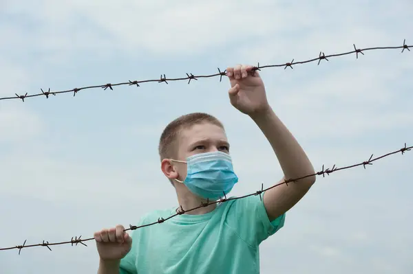 Tıbbi maskeli çocuk, arka plandaki dikenli tellere tutunuyor.