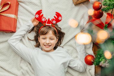 Noel Baba şapkalı mutlu bir kız Noel ağacının yanında yatıyor ve gülümsüyor. Çocuklar yeni yılı ve Noel 'i kutlamaya hazırlar. Tatil için hazırlanıyorum..