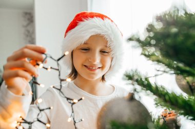 Noel Baba şapkalı çekici kız evdeki Noel ağacını ışıklarla süslüyor. Yeni yıl ve Noel kutlamalarına hazırlanıyorum..