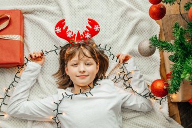 Mutlu bir kız, yeni yıl süslemeleri ve çelenk ile Noel ağacının yanında beyaz bir ekoseye uzanır. Çocuklar Noel 'i ve yeni yılı bekliyor.. 