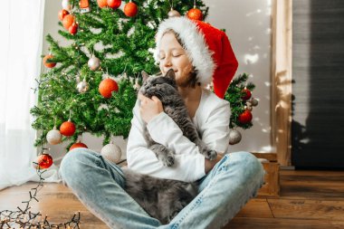 Noel Baba şapkalı bir kız Noel arifesinde Noel ağacının yanında gri kedisine sarılır. komik hayvanlar.