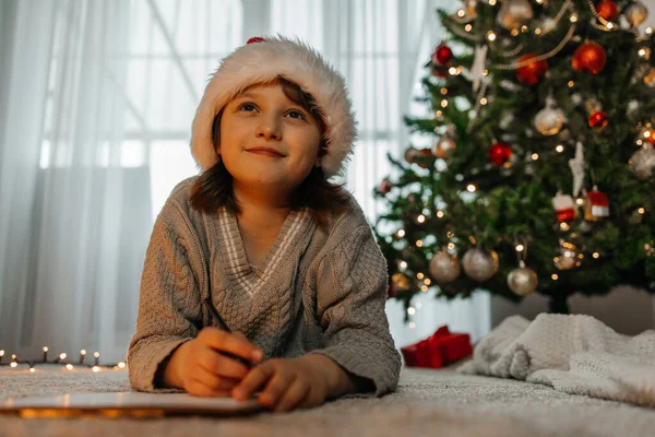 stock image A girl in a red santa claus hat writes a letter to Santa Claus on the background of the Christmas tree. 