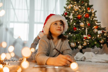 Yılbaşı ve Noel. Noel Baba şapkalı genç bir kız Noel Baba 'ya çelenk ve ışıklarla süslenmiş bir Noel ağacının yanına bir mektup yazar..