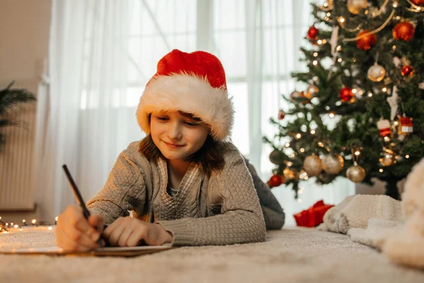 stock image New Years and Christmas. A teenage girl in a santa claus hat writes a letter to Santa Claus next to a decorated Christmas tree in garlands and lights.