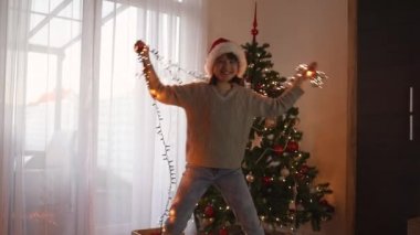 Neşeli bir genç kız Noel ağacının yanındaki bir odada yeni yıl çelengiyle dans ediyor. Mutlu Noeller ve yeni yıl.