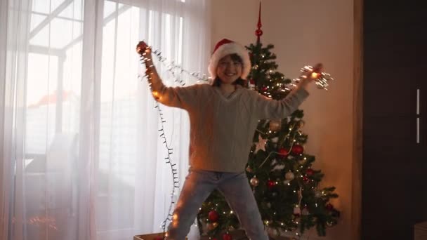 一个快乐的少女和一个新年花环在圣诞树旁的房间里跳舞 圣诞快乐 新年快乐 — 图库视频影像