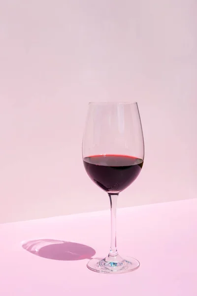 Wine glass with red wine on a pink background in the rays of the bright sun. Place for text.