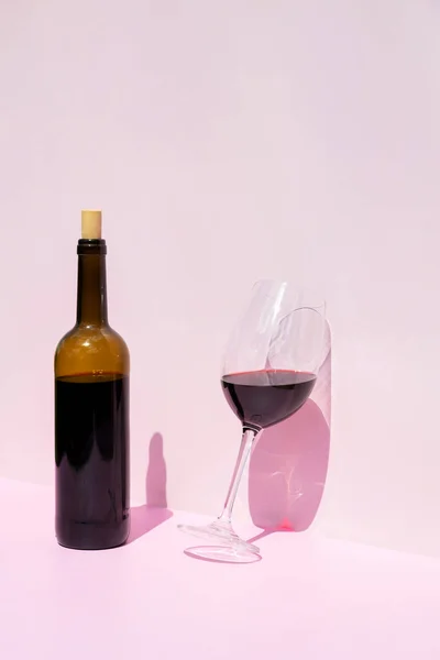 A bottle of red wine and a filled glass next to it in the sun on a pink background. Place for text.