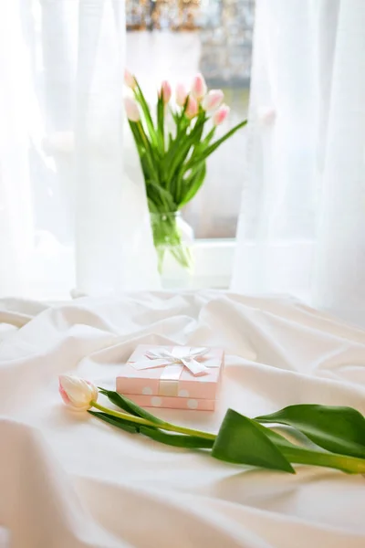 A gift box with a tulip flower on the bed and a bouquet of flowers on the window. Nice morning moments. Gifts for the holidays.